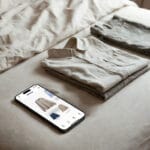 Smartphone sobre una cama con la aplicación estilo Whering mostrando un clóset digital y ropa doblada, simbolizando la organización para ahorrar dinero.