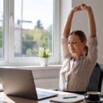 Profesional tomando una pausa activa y estirándose frente al portátil en un espacio de trabajo iluminado, como parte de un hábito saludable para prevenir el agotamiento digital.